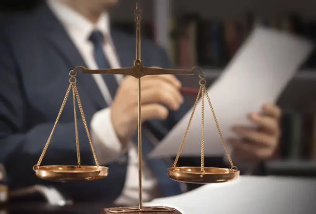 Scales of justice in foreground, lawyer in suit reading legal document in blurred background.
